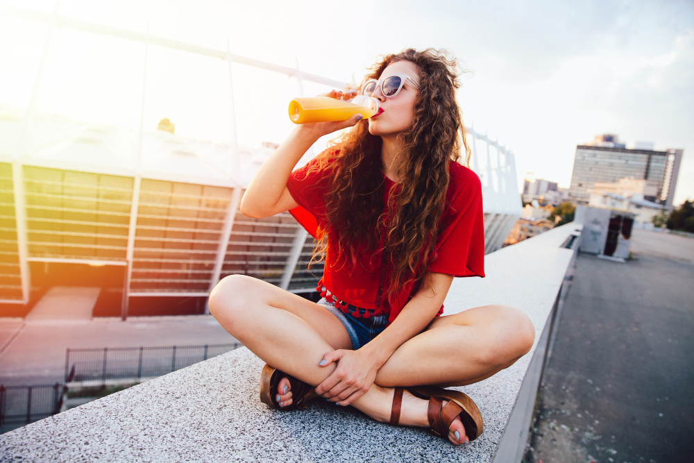 young hipster woman with curly hair sunglasses drinking fresh orange juice from bottle